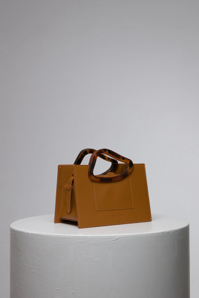 Rectangle shaped champagne calf leather saddle bag with natural resin handle holder and remowable calf leather handle strap on a white stand
