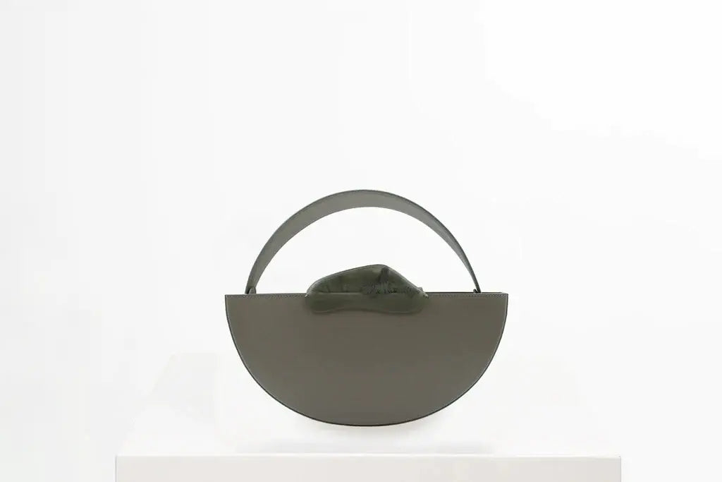 Flat topped oval structured taupe calf leather hand bag with two natural resin round handles and a removable calf leather strap on a white stand
